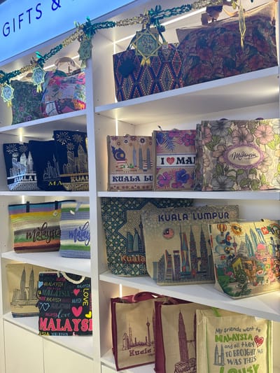 The image displays a retail store's product display in Sepang, Malaysia, featuring numerous souvenir tote bags and handbags arranged on five white, illuminated shelves. Green and gold tinsel garlands with star-shaped *ketupat*-inspired decorations are strung above the top shelf.

The top shelf showcases several large bags, including a dark green floral patterned bag, a multi-colored patchwork fabric bag, a dark bag with a red and blue geometric diamond pattern, and a large dark bag with a dense pattern of red and green hibiscus flowers.

The second shelf from the top features a row of mid-sized tote bags. On the left, three black bags depict silver or gold outlines of the Kuala Lumpur skyline and text such as "KUALA LUMPUR" or "MALAYSIA." To their right, three lighter-colored bags are visible: one with a floral pattern and "KUALA LUMPUR" skyline, another with "I ❤️ MALAYSIA" and flowers, and a third with a floral background and "Malaysia" in script.

The third shelf from the top presents two crocheted or woven bags with horizontal stripes in various colors (e.g., green, brown, blue) and "Malaysia" in script. To their right are three rectangular bags, likely jute or canvas; one with a blue geometric pattern background and a colorful Kuala Lumpur skyline, another with a beige background featuring the Malaysian flag and skyline, and a third with a vibrant, cartoon-style illustration of the Kuala Lumpur skyline and other Malaysian elements.

The bottom visible shelf displays two beige bags with outlines of the Petronas Twin Towers, one marked "Kuala Lumpur." Next to these is a black bag patterned with "MALAYSIA" in various fonts and colors, interspersed with heart symbols. Further right, two more beige bags are visible: one with a burgundy trim and a tower outline, and another with green trim featuring text that begins "My friends went to MALAYSIA and all they brought me was this..."
