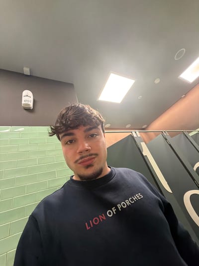 The image displays a light-skinned male with dark brown hair, a mustache, and a goatee, positioned centrally and looking directly at the camera. He is wearing a dark blue crewneck sweatshirt with the words "LION OF PORCHES" prominently displayed in white lettering on the chest, with "LION" potentially in red.
The setting is a public restroom, identifiable by its distinct features. To the subject's left, the wall consists of light green subway-style tiles on the lower section and a solid dark grey/brown painted surface above. A white Jofel-branded wall-mounted dispenser, likely a hand dryer or air freshener, is affixed to the upper dark wall. To the subject's right, a series of dark grey partitions, characteristic of toilet stalls, are visible, with partially visible white markings (appearing as parts of numbers or letters) on their surfaces. Beyond the partitions, a wall section is painted in a reddish-brown or terracotta hue. The ceiling is grey and features two large, illuminated rectangular recessed light panels, along with several smaller, circular recessed lights or sensors.
Based on the brand displayed on the subject's clothing ("LION OF PORCHES," a Portuguese brand) and the generic modern public restroom aesthetic, the location is assessed to be a public restroom within a "Lion of Porches" retail store or an associated commercial establishment in Penafiel, Portugal.