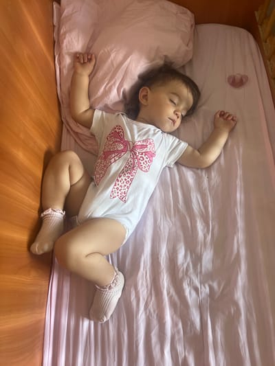 A toddler is sleeping on a pink sheet in a crib. The toddler is wearing a white onesie with a pink bow pattern and pink socks. A pink pacifier is on the sheet near the toddler's head. The crib has a wooden frame. The flooring is made of wooden planks. The scene appears to be indoors during the day.
