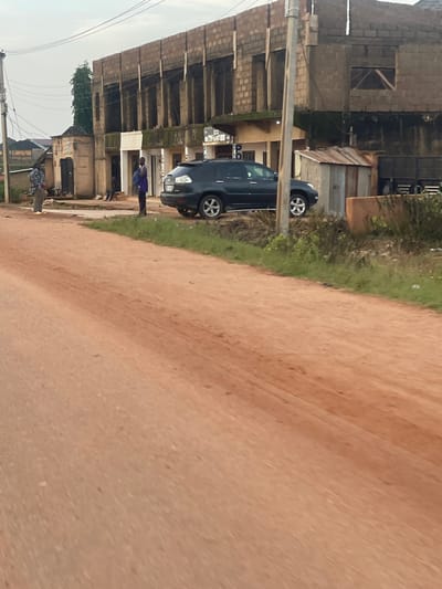 A dark blue car is parked on a dirt road next to a grassy area and some buildings. A person in a purple shirt and dark pants is standing near the car, looking towards the buildings. In the background, there are unfinished buildings made of concrete blocks, some with corrugated metal roofs. The sky appears overcast. The scene is likely taking place in a developing area, possibly a town or village in Kwang, Nigeria. The lighting suggests it could be late afternoon or early morning, given the muted light. The overall impression is one of a typical day in a community with ongoing construction and everyday life unfolding. There is no visible text in the image.