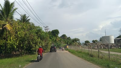 The image depicts a road scene in Blang Mee, Indonesia. A person wearing a red shirt and helmet is riding a black motorcycle on the left side of the asphalt road. A dark SUV is ahead of the motorcycle on the right side of the road. Further ahead, another motorcycle and a yellow truck are visible. The road is flanked by lush green vegetation and palm trees on the left, and a chain-link fence with barbed wire on top separates the road from a property with large rocks and a white cylindrical structure on the right. Power lines are strung across the sky above the scene, and the sky is cloudy. The overall impression is a rural or semi-rural road in Indonesia on an overcast day.