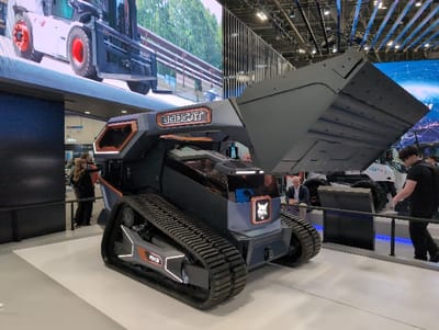 A modern, gray and black Bobcat construction vehicle with elevated bucket is on display. It features tracks and illuminated accents. A large screen behind the machine displays construction equipment. People are visible in the background, including a man in a suit and a person in a black shirt. The scene appears to be at a trade show or exhibition.