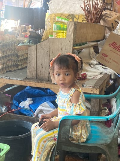 A young girl with dark hair pulled into two pigtails, adorned with light orange bows, sits on a weathered, teal plastic chair. She wears a colorful dress with a floral pattern in yellow, blue, and white, featuring ruffled sleeves. Her face is turned towards the viewer, with a tear visible on her left cheek. She holds a black object, possibly a phone, in her hands. The background suggests a market stall in Ta', Indonesia. Piles of goods are visible, including stacks of egg cartons, a large yellow sack, bottles of cooking oil, and wooden crates. A large black bucket is positioned to the left of the chair, containing dark liquid. The overall environment appears cluttered and somewhat disorganized, indicative of a busy marketplace.