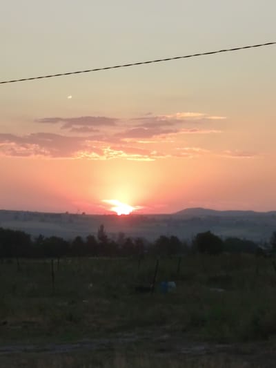 This is a vertical, medium-shot photograph of a rural landscape during sunset. The sky is a gradient of pale yellow at the top, transitioning to a vibrant orange and pink as it meets the horizon. Wispy, pink-tinged clouds are scattered across the upper half of the sky. A thin, dark utility wire is stretched horizontally across the upper portion of the image.
The sun is setting behind a line of distant hills, its bright rays fanning out in a dramatic display, casting a warm glow over the scene. Below the sky, a dark, silhouetted treeline forms a gentle curve along the horizon. In the foreground, a field of dry, grassy terrain is visible. Several dark, vertical fence posts are scattered across the field. The lower portion of the image is darker, suggesting the approaching twilight.
There is no discernible text in the image. The overall mood is peaceful and serene, capturing the beauty of a sunset in a natural, undeveloped area. The setting appears to be a countryside location, possibly agricultural land, given the field and fence posts. The time of day is clearly sunset, with the warm colors and fading light. The weather appears clear, allowing for an unobstructed view of the sunset.