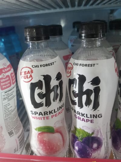 The image displays a close-up, eye-level view of several chilled "Chi Forest" brand sparkling beverage bottles, positioned within a refrigerated display unit. In the foreground, two clear plastic bottles with black ribbed screw caps are prominent. The bottle on the left is "Sparkling White Peach" flavor, featuring a white label with "BEBAS GULA" (Sugar-Free) in a red circle, the brand name "CHI FOREST™", a large stylized black "Chi" logo, and an illustration of a white peach with a green leaf at the bottom. The bottle on the right is "Sparkling Grape" flavor, with an identical label design but depicting purple grapes and green leaves. Both bottles appear to have condensation on their surfaces.

In the blurred background, additional bottles of the same brand and others are partially visible. To the far left, a portion of another "Chi Forest" bottle displays nutritional information in Indonesian or Malay, including "O Kalori" (0 Calories) and "INFORMASI NILAI GIZI" (Nutritional Information). A blue bottle is also discernible behind the front left bottle. The immediate environment includes a metal wire shelf supporting the bottles, and the ribbed white interior ceiling or upper shelf of the refrigerator above them. The scene is well-lit, indicative of internal refrigeration lighting. No people are visible, nor are any actions or interactions depicted. The specific location within Naung Bo Oe, Myanmar, is not discernible from the image content; the visible product labeling suggests a region where Indonesian or Malay is spoken.