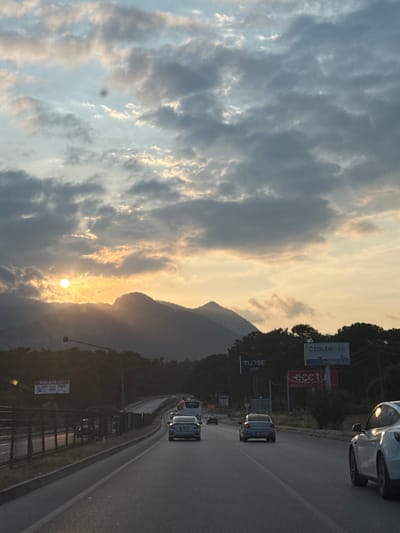 The image captures a multi-lane highway scene at sunset in Döşemealtı, Turkey. The sky is largely covered by stratocumulus clouds, illuminated by the setting sun, which appears as a bright, distinct yellow-orange disc positioned low on the horizon on the left, partially obscured by the dark silhouette of mountains. The western sky exhibits a gradient of warm yellow, orange, and light blue hues, transitioning to darker grey clouds overhead. A prominent dark, circular blemish, consistent with sensor dust, is visible in the upper central portion of the sky.
The asphalt road, running from the foreground into the distance, accommodates vehicular traffic. A white Tesla Model Y is visible in the right foreground, angled away from the viewer, with intense sunlight reflecting off its side panel. Further along the road, in the central lanes, a dark-colored sedan (appearing to be a Honda Civic) is prominent, followed by additional less distinct vehicles including another sedan and a partial view of a white bus on the far left lane.
Along the right side of the road, a series of large advertising billboards are erected. One billboard clearly displays "ÖZDİLEKPARK ANTALYA." Below it and to the left, another sign reads "TURSE," and a red billboard partially shows "ece tu." On the left side of the road, beyond a metal barrier, a billboard for "anpa gross" is visible. Dense green trees and foliage line both sides of the highway, extending into the background and covering the lower slopes of the silhouetted mountains. Overhead gantries and traffic signal structures are also present along the roadway. The ambient lighting indicates dusk or late afternoon.