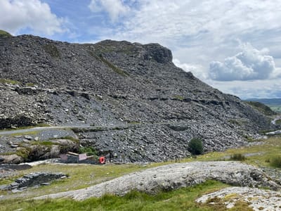 The image depicts a vast, rocky mountainside in Tanygrisiau, United Kingdom. The terrain is dominated by loose stones and slate, forming a steep incline. A winding road snakes its way across the lower part of the mountainside. In the foreground, a body of water flows through a rocky channel. There are a few safety barriers and a lifebuoy near the water, indicating a potential recreational area. A sign is visible which reads "DIM NÔS SWIMMING". Several people are present, including one individual standing by the water and a couple of distant figures on the road. The sky above is a mix of blue and white clouds, suggesting a partly cloudy day. The overall environment appears rugged and natural, with sparse vegetation in some areas.