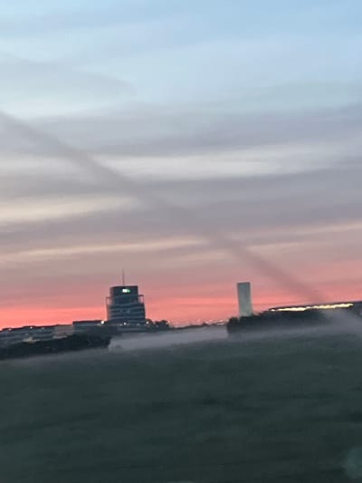 The image captures a hazy sunset over a semi-urban landscape in Leudelange, Luxembourg. In the mid-ground, a distinctive cylindrical building with a rounded top, identified as the "Centre de Recherche Public Henri Tudor," stands tall, illuminated by a green light near its summit. To its right, a slender, rectangular tower is visible, casting a long shadow. A line of lower buildings stretches across the horizon, with some windows reflecting the warm hues of the sunset. The foreground consists of a dark, grassy field, partially obscured by a misty haze that seems to emanate from some unseen source, possibly a sprinkler system. The sky above transitions from a soft blue at the top to a vibrant pink and orange gradient near the horizon, with subtle cloud formations creating a layered effect. The overall atmosphere is serene, with the light pollution from the buildings and the hazy conditions contributing to the diffused light.