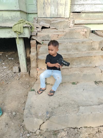 A young boy sits on concrete steps leading to a weathered, wooden structure. He wears a black shirt, light blue jeans with a ripped knee, and sandals. He holds a toy gun in his hands. The steps and surrounding area show signs of age and disrepair. A green cloth hangs from a support beam of the structure. The background features a mixture of textures, including cracked concrete, peeling paint, and exposed brick.