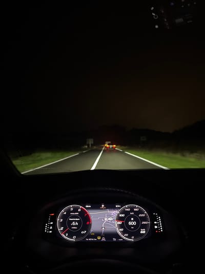 The image depicts a first-person view from the driver's seat of a modern vehicle traveling at night on a two-lane road in Kaysersberg-Vignoble, France. The digital instrument cluster is prominently displayed, showing the time as 06:44, an outside temperature of 12.5°C, and the vehicle's speed at 68 km/h. Other dashboard details include a trip odometer reading 4214.1 km, average fuel consumption of 6.4 l/100km since departure, and a visible tire pressure warning light. The road ahead is illuminated by the vehicle's headlights, revealing a wet asphalt surface with white lane markings and grassy shoulders on both sides. The red taillights of another vehicle are visible in the distance, traveling in the same direction. The environment surrounding the road is dark, with indistinct vegetation silhouettes against the dark sky. Two faint rectangular road signs are visible on the left side of the road ahead.