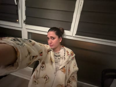 A woman in a light-colored, patterned onesie stands indoors. She also wears a white t-shirt with writing. Her dark hair is pulled back in a bun. Behind her is a wall with horizontal paneling and windows with white frames. The image is rotated so the woman appears upside down.