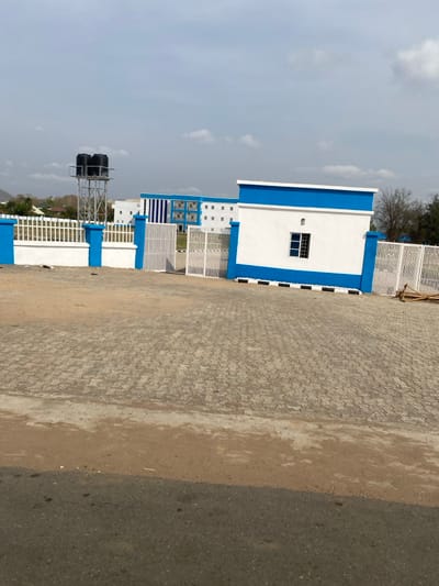 This image depicts the entrance and interior of a facility located in Abobo, Nigeria, captured during daylight hours under a partly cloudy sky.
The foreground consists of a dark grey asphalt road at the bottom, transitioning to a strip of reddish-brown dirt, then to a large expanse of grey interlocking pavers.
The midground features a perimeter wall and gate system. A white concrete wall with blue painted trim and pillars runs horizontally. To the left, a white metal fence extends from a blue pillar. Centrally, a wide white metal gate, composed of multiple vertical bars, is partially open. To the right of the gate is a small, white rectangular building, possibly a guardhouse, with blue top and bottom trim and a single barred window. Further right, more white metal fencing is visible, with some wooden planks or construction debris stacked near the fence line on the far right.
In the background, beyond the gate, multiple multi-story buildings are visible. These structures are predominantly white with prominent blue vertical stripes and trim, featuring multiple windows. To the left, a tall metal water tower supports two large black cylindrical water tanks. Distant hills or mountains are faintly visible on the far left horizon. The sky is light blue with scattered white cumulus clouds.
No individuals are visible, and no discernible actions or interactions are occurring. The scene appears static.