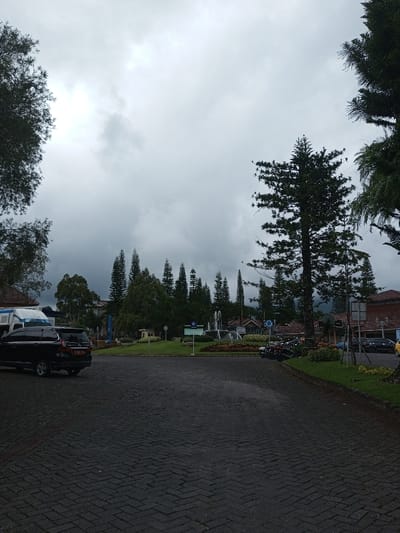 A paved parking area leads to a roundabout with trees, a fountain, and a sign. A black SUV is parked on the left, near a white van. Motorbikes are parked on the right. Tall evergreen trees frame the scene. The sky is overcast and gray. Buildings are visible in the background, partially obscured by trees. A patch of green grass is visible on the right side of the path.