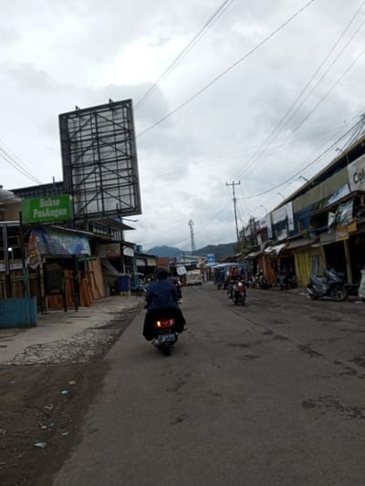 The image depicts an urban street scene in Melakudikadu, India, under an overcast sky. A paved road, showing signs of wear and scattered debris, extends from the foreground into the distance. In the immediate foreground, a person riding a dark scooter, wearing a dark jacket and a dark garment over their legs, drives away from the viewer, with the scooter's taillights illuminated. Further down the road, several other motorbikes are visible, with at least two carrying passengers, moving towards or away from the viewer.
Buildings line both sides of the street. On the left, a multi-story structure features a large, empty metal billboard frame atop its roof. Below the billboard, a prominent green sign with white text reads "Bakso Pasangan". Adjacent to this building, other structures, possibly shops, are visible, with wooden planks stacked near the sidewalk. On the right side of the road, a continuous row of single-story buildings, appearing to be shops or residences, extends into the background. Some of these structures have various facades and awnings, and a partial sign displaying "Colom..." is visible on one.
Overhead utility lines crisscross the sky, supported by wooden and metal poles along the street. In the far distance, a cellular tower or antenna structure stands against a backdrop of rolling hills or mountains beneath the cloudy horizon.