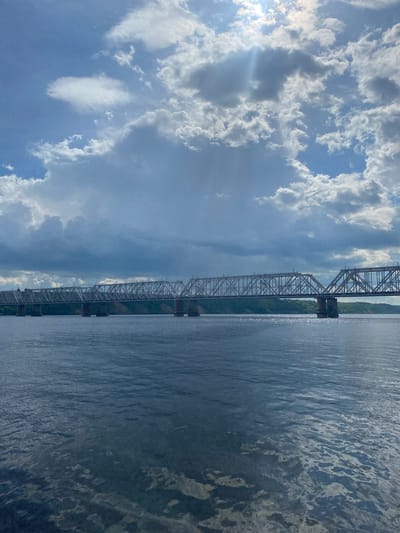 This media file depicts a wide, calm body of water alongside a large, steel truss bridge. The bridge spans a considerable distance, supported by multiple piers. On the left side of the frame, a dramatic sky is visible, filled with a mix of fluffy white clouds and darker, more ominous storm clouds. Sunlight breaks through in some areas, casting rays and creating lens flares. The water reflects the sky and the bridge, with subtle ripples disturbing the surface. Greenery lines the far bank of the water, partially obscured by the bridge's structure.

The scene appears to be captured during the daytime, given the presence of sunlight and the visible clouds. The weather seems dynamic, with both sunny patches and developing storm clouds. The presence of the large bridge suggests an industrialized or transportation-focused location. Without further information or visible text, a precise location cannot be determined, but the style of the bridge and the landscape could be consistent with a river setting. No people are visible in the media file. The overall mood is one of natural grandeur and perhaps a touch of atmospheric tension due to the weather.