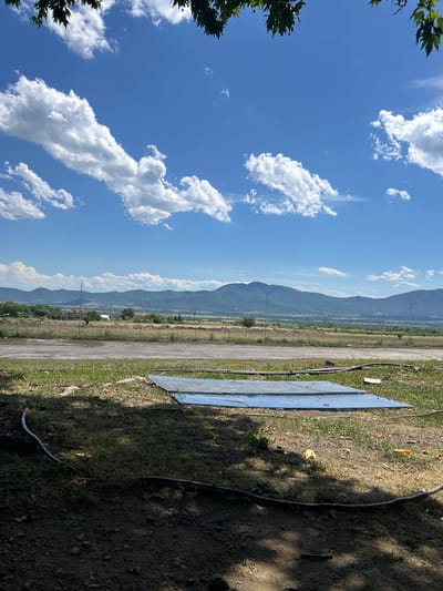 The image shows a rural landscape with mountains in the background under a blue sky dotted with white clouds. In the foreground, there is dry grass, dirt, and some scattered debris, including a blue tarp or sheet. A coiled hose or cable runs along the ground. Beyond the immediate foreground, there is a dirt or gravel road, followed by more dry grass and a fence. Further in the distance, there are scattered buildings, trees, and the aforementioned mountains. The scene suggests an outdoor, possibly undeveloped or construction site area near Sopot, Bulgaria.
