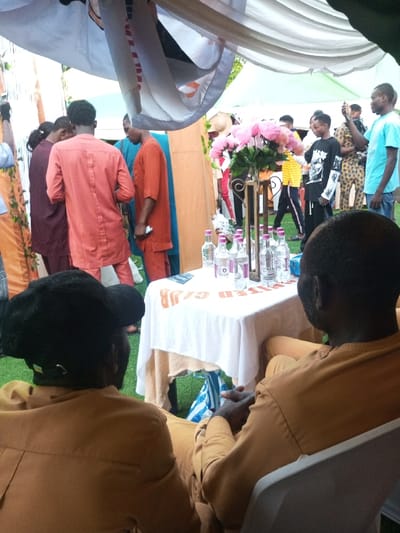 The image depicts an outdoor gathering in Amokvo, Nigeria. In the foreground, two individuals are seated with their backs to the viewer in white plastic chairs. Both wear matching orange-brown long-sleeved tunics and trousers; the person on the left also wears a black baseball cap. A white-covered table positioned in front of them holds approximately eight small clear bottles with pink caps, a decorative centerpiece featuring pink artificial flowers on a dark stand, and a white towel with red lettering, reading 'UNITED CLUB' in reverse orientation. The ground beneath is covered in green artificial turf.
The midground reveals a crowded scene of standing adults under a white draped canopy. Observable individuals include a person in a brick-red long tunic, another in an orange short-sleeved tunic, and an individual in a bright blue t-shirt actively holding a smartphone towards the right. Other attendees wear various garments, including a dark red outfit, a black t-shirt with white graphics, a yellow and black striped shirt, and patterned fabrics. The background shows additional white and green canopy structures and more indistinct figures.