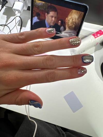 The image presents a hand with decorated fingernails. The index finger nail is gold-colored with a textured, studded design. The middle finger nail is primarily silver with a blue border. The ring finger has a semi-transparent base with a silver swirl design. The pinky finger has a metallic pink-silver color. The thumb has a blue marbled design with white or cream accents.

The hand is positioned over a table surface with multiple items on it. There is a tablet displaying a person in a suit. Beside the tablet are power adapters and cables. A white container of what appears to be cotton rounds is at the top of the frame. A white, rectangular card is on the white table surface. A portion of a grey sweatshirt is visible on the right side.

There is insufficient information in the image to determine the location within the city of Hull, United Kingdom.