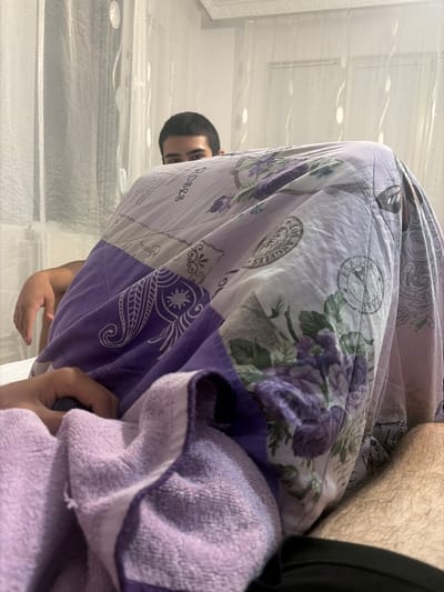 In a domestic indoor setting, likely a bedroom in Novi Iskar, Bulgaria, a person is partially visible behind a purple and white patterned duvet adorned with floral designs and what appear to be postage stamp motifs. The individual's face, specifically their eyes and forehead, is peeking out from under the duvet. Their dark hair is short and neat. To the left of the frame, another person's arm and hand are visible, resting on a folded light purple towel. The background features sheer white curtains, suggesting a window. The overall impression is one of casual relaxation or a playful moment within a home.