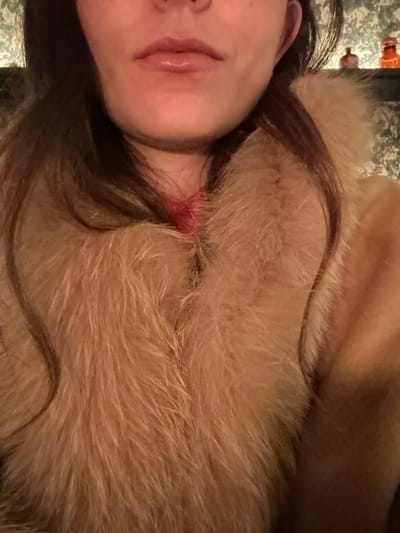 A woman's face, including part of her nose, mouth, and ear, is visible. She has dark hair. She wears a tan-colored fur garment. A red, irritated area is visible on her neck. A surface with a patterned design is in the background. The image appears to be taken at an angle.