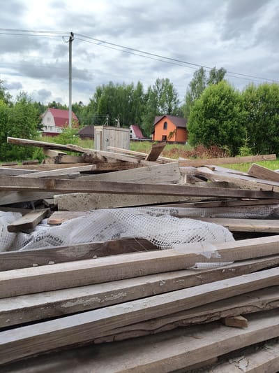 This media file depicts a pile of discarded wooden planks in an outdoor setting, likely a construction or renovation site. The planks are of various sizes and appear weathered, with some showing signs of decay and peeling paint. A white, mesh-like material is partially visible underneath some of the planks, possibly a protective covering.
In the background, several houses are visible, suggesting a semi-rural or suburban location. One house has a prominent red roof, while another is painted a bright orange. Lush green trees and bushes surround the structures, indicating a season with active foliage, likely summer or late spring. A utility pole with overhead wires stands tall in the middle ground, further contextualizing the environment.
The sky is overcast with heavy, grey clouds, suggesting a cloudy or impending rainy day. The overall lighting is diffused, and there are no strong shadows. The scene appears still, with no visible people or animals. The focus is primarily on the pile of wood in the foreground, emphasizing its disarray.
Some text is visible on the white mesh material. Though partially obscured and blurry, it appears to be in Cyrillic script. Phrases like "СТОРОНА" (meaning "side") and other partial words like "ЯКО" can be discerned. The location context provided is Скурыгино, Russia.