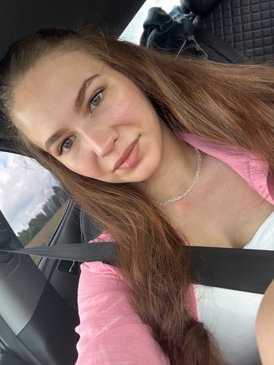 A young woman with long, brown hair and green eyes is shown in a selfie taken from inside a car. She is wearing a pink jacket over a white top and a delicate, sparkly necklace. The seatbelt is visible across her chest. Through the car window, a blurry view of trees and a cloudy sky can be seen. The background inside the car includes a black quilted seat and a dark fabric draped over it.