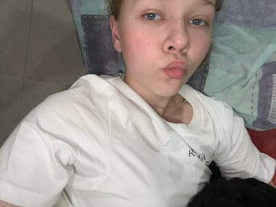 A young Caucasian woman with blonde hair and blue eyes is lying down and looking at the camera with her lips puckered. She is wearing a white t-shirt. She is on a couch or bed with pillows behind her. The background is blurred and consists of a patterned fabric. There is no information in the image to indicate the location in the city of Klepiki, Russia.