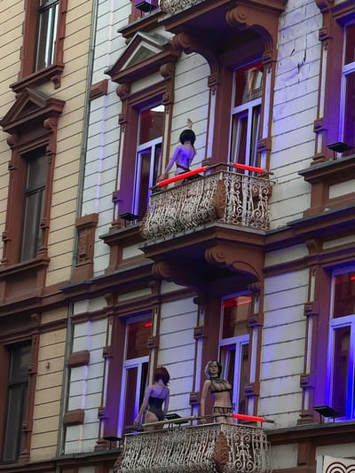 The image shows a multi-story building in Frankfurt, Germany, with several mannequins displayed on balconies. The building has a traditional architectural style with red-brown and cream-colored facades, ornate balconies, and large windows. The mannequins are dressed in revealing attire, including lingerie and swimsuits. Some of the balconies are illuminated with colored lights, creating a vibrant and somewhat provocative atmosphere. The scene appears to be set in an area that caters to adult entertainment or has a similar commercial focus.