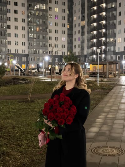 The image depicts an outdoor nighttime scene in Новогорелово, Russia, within the courtyard of a residential apartment complex. A female subject, appearing to be in her twenties or early thirties with blonde, wavy hair, stands in the foreground, slightly right of center. She is wearing a dark, long overcoat over a red top and holds a large bouquet of approximately 25-30 red roses with green foliage and a pink ribbon. She looks upwards and to her left with a slight smile. The ground consists of a paved pedestrian path on the right, featuring a circular metal manhole cover, and grassy areas with bare earth patches and sparse, leafless trees. In the background, two multi-story apartment buildings with numerous illuminated windows dominate the frame, suggesting a populated residential area. Streetlights provide artificial illumination throughout the courtyard. In the mid-ground, a children's playground with a slide is visible on the left, and a wooden gazebo or pergola structure is present on the right. Several indistinct human figures can be observed near the gazebo, and at least one parked vehicle is visible near the playground structures.