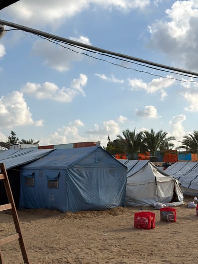 The image captures an outdoor scene in a-Zawayda, Palestinian Territory, featuring a temporary encampment under a bright blue sky with scattered white cumulus clouds. The foreground consists of sandy, light brown ground. On the left, a portion of a brown wooden ladder is visible. In the mid-right, several red bags or containers are present on the sand. The midground is occupied by multiple tents. A prominent, gabled-roof blue tent stands in the center-left, displaying two rectangular window flaps and partially visible text, including "UNRWA", on its fabric. To its right, a row of white or light-colored gabled-roof tents extends across the frame; one of these tents appears slightly ajar at its entrance. In the background, numerous green palm trees are visible, behind which lie structures with distinct colored panels, including orange, red, and blue. A thick, horizontal metal pipe or beam spans the upper portion of the image, with thin black wires running beneath it. No individuals are depicted in the scene.