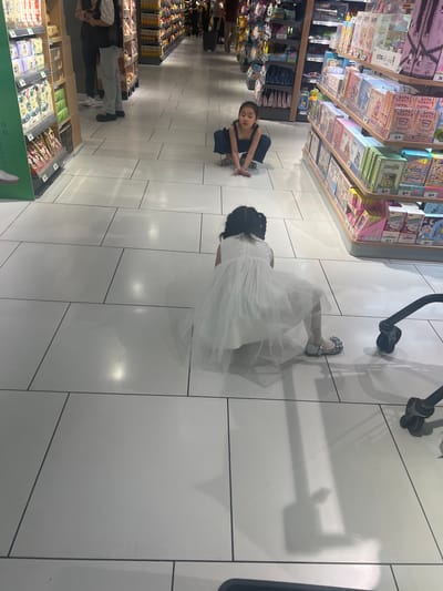 In the Liandu District, China, two young girls are in a grocery store aisle. One girl, wearing a dark blue jumpsuit, is crouched on the floor, looking forward. The second girl, wearing a white dress and silver shoes, is also on the floor, facing away from the camera, with her legs bent and her arms resting on the ground. In the background, shelves are stocked with various products. A shopping cart is partially visible on the right side of the frame. The floor is tiled with large, white tiles.