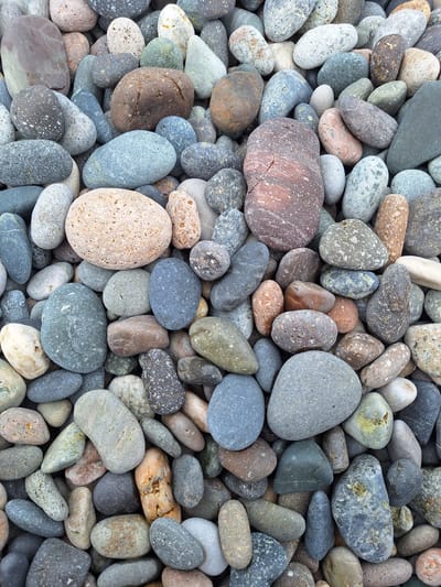 This image is a close-up, overhead view of a collection of smooth, rounded pebbles. The pebbles vary in size, shape, and color, with hues ranging from light gray, beige, and white to darker grays, blues, and some with reddish-brown and green speckles. They are densely packed together, creating a textured surface. There are no people or discernible activities taking place. The lighting appears to be natural and even, suggesting it could be daytime under an overcast sky, or in diffused light. The setting is clearly a beach or shoreline, given the appearance of the stones. There is no visible text in the image. The overall impression is one of natural, geological formation and the tactile quality of the pebbles.