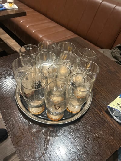 A silver tray sits atop a dark wooden table, holding ten empty Guinness glasses. The glasses are arranged in a neat, triangular formation. Each glass bears the Guinness logo. The table has a worn, textured surface. Part of a tan-colored leather bench is visible on the left side of the frame.