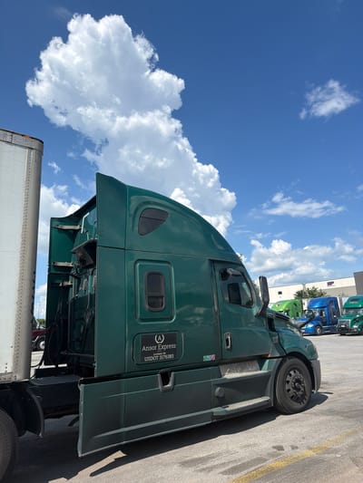 The image shows a row of semi-trucks parked in an outdoor lot under a partly cloudy blue sky. The main focus is on a dark green semi-truck in the foreground, seen from its right side. This truck has the logo "Ansor Express LLC" with "Loveland, OH" and "USDOT 3176383" printed on a black rectangular decal on its side. A portion of a white trailer is visible to the left of the green truck. In the background, other semi-trucks of various colors, including blue and bright green, are parked. A light-colored building with trees in front of it is visible in the far background. The ground is asphalt with yellow painted lines. There are no people visible in the image. The location is the city of Lebanon, United States of America.