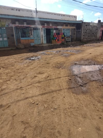 The image depicts an unpaved dirt road in Njoro ward, Kenya, with commercial structures lining one side. The road surface is brown earth, marked by multiple visible puddles of standing muddy water and damp patches, indicating recent moisture. Shadows are cast across the road from an unseen source to the left.
On the right side of the road stands a row of single-story buildings constructed from unpainted concrete blocks with a corrugated iron roof. The first visible establishment from the left is labeled "LIFETIME SALON & CURTAINS," featuring a light blue, partially open metal door and an orange counter or display stand in front. Adjacent to this, another shop has an open entrance with a vertically displayed sign that reads "M-PESA" and other partially obscured text, including "NEO-DILIMIGI" or similar, indicating a mobile money service point. Outside this shop, a large, organized stack of brightly colored gas cylinders (pink, orange, yellow, green, and light blue) is visible. A bicycle with a basket is parked next to these cylinders, near a small, obscured sign. Further to the right, another section of the building presents a closed light blue metal door, followed by a grey concrete block wall.
The sky above is partly cloudy with patches of blue. Overhead power lines are visible against the sky. No individuals are present in the scene, and no specific actions or interactions are depicted. A large stone rests on the ground near the "LIFETIME SALON."