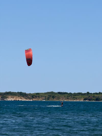 A single individual is kitesurfing on a body of water near Atiya, Bulgaria. The person is wearing dark clothing and is standing on a board, being pulled by a large, red kite suspended in the clear blue sky. The kite has some yellow and black markings on it. The water is a deep blue with moderate ripples. In the background, a tree-covered coastline with some rocky outcrops and a sandy beach with scattered umbrellas is visible. A white sailboat is anchored offshore, and a few other small boats can be seen in the distance. The overall scene depicts a sunny day with good conditions for watersports.