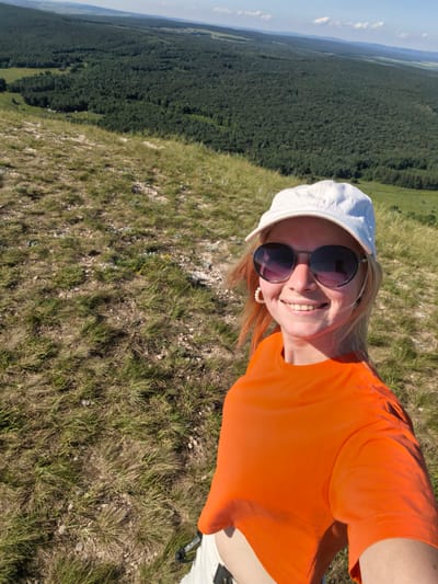 A young woman is taking a selfie on a grassy hill overlooking a vast, dense forest. She is wearing a white baseball cap, oversized round sunglasses, and an orange t-shirt. Her blonde hair with orange streaks is visible around her face. The background shows a panoramic view of rolling hills covered in a thick, dark green forest that stretches to the horizon. The sky is blue with a few scattered white clouds. The foreground is a dry, grassy slope with patches of rocks and small yellow flowers. The woman is smiling and appears to be enjoying the scenic view. The location is the city of Shikhan, Russia.