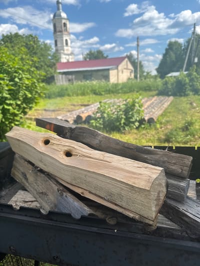 In the foreground, several logs are stacked within a dark metal structure, possibly a grill or fire pit. The logs vary in size and color, with some appearing freshly cut and light brown, while others are darker and weathered. One of the lighter logs has two distinct circular holes drilled into its surface. Behind the logs, the scene opens up to a grassy outdoor area. In the middle ground, rows of cut logs are laid out on the grass. In the background, a building with a red roof stands, and to its left, a tall, white bell tower with a gray dome dominates the skyline under a bright blue sky with scattered white clouds. Green trees and foliage are visible throughout the scene, suggesting a rural or semi-rural environment. The overall impression is of a scene set in the countryside, possibly in or near a village, with evidence of preparation for a fire or outdoor cooking.