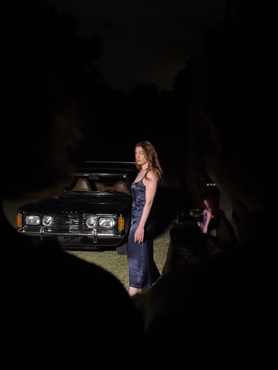 A woman in a sleek, dark blue dress reclines on the hood of a black vehicle, illuminated by headlights. The scene is shrouded in darkness, with only the front of the car and the woman visible. The background features grass. Above, several figures are visible, holding up phones to take pictures. The light creates stark contrasts between light and shadow.