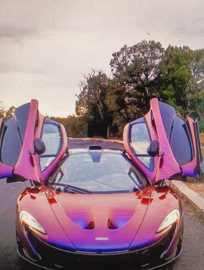 A front-facing, medium shot captures a red and purple McLaren P1 supercar with its dihedral doors open. The car is parked on an asphalt surface, with a paved border and a dirt shoulder visible to the right. Behind the car, a line of trees and vegetation borders a wooded area. The sky is overcast, with a bright gradient suggesting late afternoon or early morning light. There are no people visible in the image. The location is not specified as being in Sluck, Belarus.