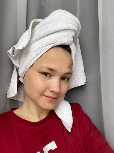 An overhead, rotated portrait shows a young person with dark hair, lying down and looking towards the viewer with a serene expression. The subject is wearing a red t-shirt and has a white, fluffy towel wrapped around their head, suggesting freshly washed hair or a spa treatment. The image is rotated 90 degrees clockwise, placing the person's head on the left and their torso extending to the right.

The person's face is central, with dark eyes looking directly at the camera, and a faint, closed-mouth smile. Their skin appears clear, with dark eyebrows. A small mole is visible on the upper chest/neck area. The red t-shirt has a white, partially visible design or text on the right side. The white towel is securely wrapped around the head, leaving the face exposed. A small white label with green text, reading "ESSENTIALS," is visible on the towel near the bottom left of the frame.

The background is simple and uncluttered, consisting of a plain gray surface where the person is resting, and a lighter, textured white surface above it, possibly a sheet or a wall. The overall impression is one of relaxation and calm. The location context is Peresvet, Russia, though no specific geographical cues are visible in the image itself.