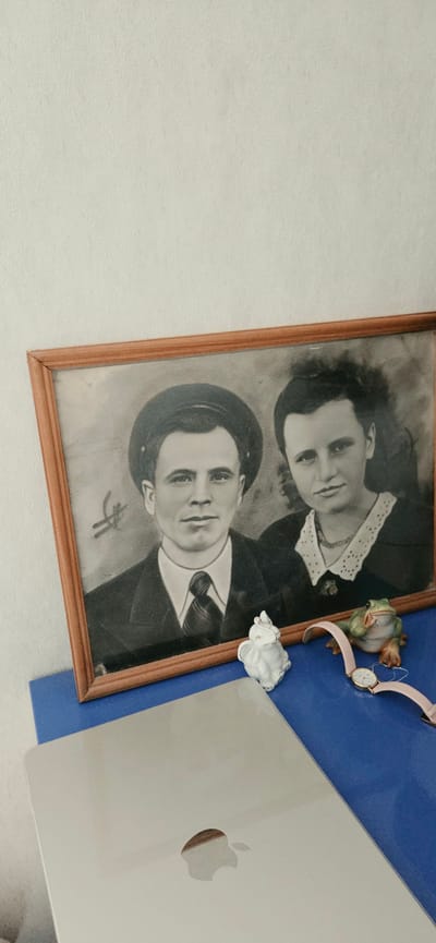 A framed black and white photograph of a young couple is displayed on a blue surface. The photograph features a man wearing a hat and suit, and a woman with dark hair and a white collar. In front of the photograph, also on the blue surface, are a small white elephant figurine, a ceramic frog figurine, and a pink wristwatch. Partially visible in the foreground is a silver laptop with the Apple logo. The background is a textured off-white wall. There is no indication of the city of Lipetsk, Russia.