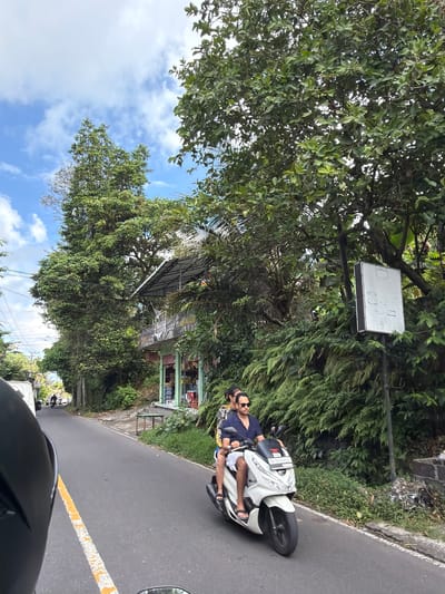 The image captures a scene on a road in Tampaksiring, Indonesia. The foreground shows the helmet of a rider, indicating a first-person perspective from a motorcycle. To the right, a white scooter is seen with two individuals riding on it. The male rider in front is wearing a navy blue shirt and white shorts, with sunglasses on. The female passenger behind him is wearing a patterned dress. They are moving along the asphalt road which has a broken yellow line marking the lane. The environment is lush and green, with tall trees and dense foliage lining the sides of the road. A small building with a storefront, possibly a shop, is visible on the right side of the road, nestled among the greenery. A white sign on a pole is also present. In the distance, the road continues, with other vehicles and more trees visible. The sky is blue with scattered white clouds.