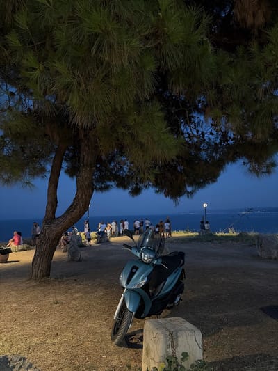 A group of people are gathered outdoors, seemingly for an evening event. The scene is set by a body of water, likely the sea, which stretches out towards a horizon dotted with distant lights, suggesting a coastal town or city. A streetlight illuminates the area, casting a warm glow on the ground, which is covered in dry grass and dirt.
In the foreground, a teal-colored scooter is parked on the right side of the frame, its headlights on. To the left, a large pine tree dominates the composition, its branches casting shadows. The tree has a thick, textured trunk and numerous pine needles, some of which are dimly lit by the artificial light.
The people, dressed in casual attire, are standing and sitting in small groups along a path or promenade that runs parallel to the water. Their relaxed postures and the gathering suggest a social occasion, perhaps watching a sunset or enjoying the evening air. The time of day appears to be twilight or early evening, with the sky a deep blue.
The overall mood is peaceful and relaxed. The location cues of the sea, the dry vegetation, and the architecture style of the scooter, combined with the provided context of Kassandra Municipal Unit, Greece, suggest a Mediterranean setting. There is no visible text in the image.