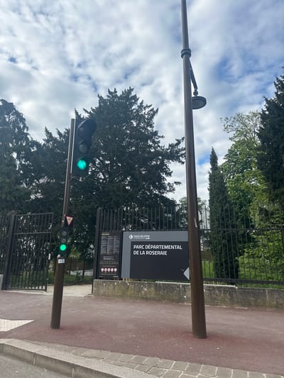 The image shows the entrance to the Parc Départemental de la Roseraie in Châtenay-Malabry, France. A sign with the park's name is visible next to a walkway. A set of traffic lights are also present, both displaying green lights. Trees are visible in the background, and the sky is cloudy.