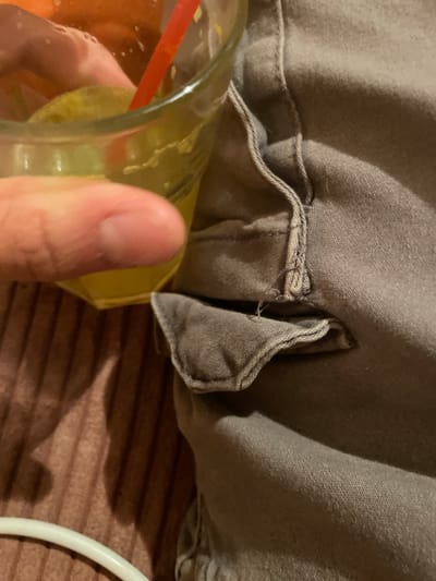 The image displays a close-up view of a person's hand holding a glass of yellow liquid, possibly a drink with ice and a red straw. The glass is partially obscuring a folded gray fabric, likely clothing, which shows stitching and some loose threads. The fabric is bunched up, creating folds and shadows. In the background, a brown textured surface, possibly a bedspread or carpet, is visible, with a white curved object, perhaps a hanger, on the right side. The lighting appears to be indoor, with a slight warm tone. The focus is on the hand, the drink, and the fabric detail. There is no visible text. The scene suggests a casual, indoor moment, possibly in a home environment. The location context provided is Malevo, Bulgaria, but there are no specific visual cues within the image to confirm this.