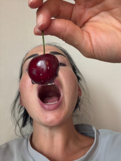 The image shows a close-up, upside-down view of a person holding a single, dark red cherry with a green stem. The cherry has water droplets on its surface. The person's mouth is open, as if about to eat the cherry. Their hand is visible in the foreground, with fingers pinching the stem. The person has dark hair and appears to have freckles. They are wearing a light-colored garment, possibly a t-shirt or sweatshirt, around their neck and shoulder area. The background is a plain, light-colored surface. The lighting suggests an indoor setting, possibly with natural light. No text is visible in the image. The location is identified as Koltushi, Russia. The activity depicted is the presentation of the cherry for consumption.