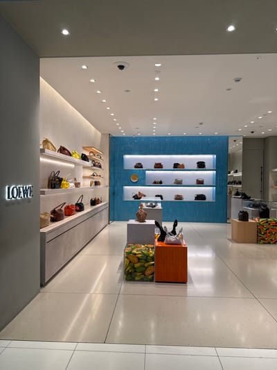 The image shows the interior of a Loewe boutique, likely located in a shopping mall in the city of Nong Prue Subdistrict, Thailand. The boutique features a minimalist design with white and gray walls, polished concrete floors, and bright, focused lighting.
The main display area showcases a variety of Loewe handbags on illuminated shelves. The bags are arranged in rows, with different styles and colors visible, including tan, burgundy, yellow, black, and red. The shelves are recessed into a light-colored textured wall, with a subtle upward glow.
To the right of the handbag displays, a section of wall is covered in vibrant blue, textured tiles, creating a striking visual contrast. More handbags are displayed on shelves within this tiled section.
In the foreground, there are display cubes. One cube is orange and contains abstract black and silver objects that resemble origami birds or bows. Another cube is clear and filled with cut papayas and avocados, adding a touch of natural color and texture to the retail environment. A further display cube to the right contains what appears to be a collection of chili peppers in various shades of green and orange.
The "LOEWE" logo is prominently displayed in illuminated lettering on the left-hand wall. The overall impression is one of luxury and modern design, typical of high-end fashion retail spaces. There are no people visible in the image.