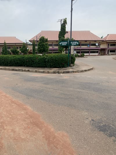 This media file depicts an outdoor scene in Wukari, Nigeria. The primary subjects are a two-story building with a pink-tiled roof and a paved road. The building features a beige facade with purple accents around the windows and has visible air conditioning units and satellite dishes on its exterior. Green, well-maintained bushes and trees line the front of the building.
A prominent object in the foreground is a street signpost with two green directional signs. One sign points left and reads "PROF. JULIUS OJOJE ROAD." Another sign, positioned below the first, also points left and contains the same text, "PROF. JULIUS OJOJE ROAD." A tall, slender lamp post is also visible on the left side of the frame.
The scene is set on an asphalt road that curves around a small, concrete-bordered island covered in greenery. The road appears to have some wear and tear, with patches of discoloration. The sky is overcast, suggesting a cloudy or potentially rainy day. The overall mood is neutral and observational. No people are visible in the frame, and no specific activity or event is taking place; it appears to be a static view of the location. The presence of the road sign indicates that this is a named street within the area.