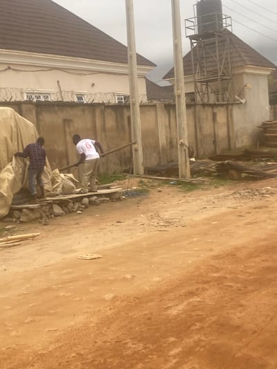 The image shows two men working on a construction site in Iddo Tudunwada, Nigeria. One man, wearing a checkered shirt and jeans, is pulling a large, tan tarp. The other man, wearing a white t-shirt with the number 6 and tan pants, is carrying a wooden plank. They are standing on a makeshift platform made of wood and rocks. Behind them is a wall topped with barbed wire, and beyond that, buildings and a water tank. The ground is a reddish-brown dirt. The scene appears to be an outdoor construction area with materials like rebar and stacked wood visible in the background.